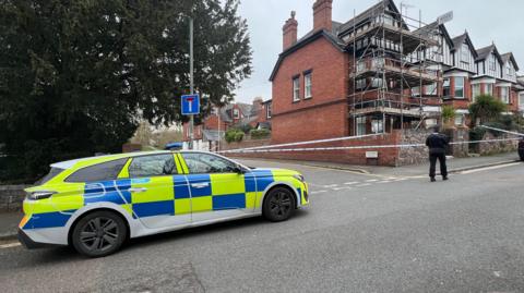 A police car is parked next to the street, there is a police cordon in place and the back of a police officer walking nearby.