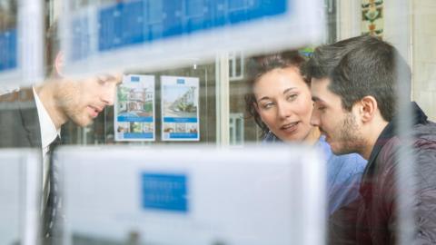 A man and woman talk to an estate agent in their office, pictured through the window.