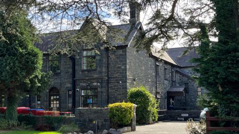 An exterior shot of the Ty Newydd bail hostel in Bangor, Gwynedd. . There are bushes and trees around the property which is a stone building. 