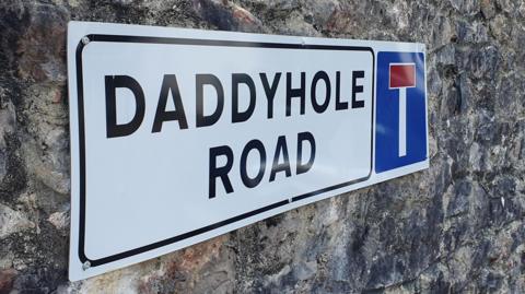 Close up of Daddyhole Road street sign. The white sign is displayed on a stone wall.