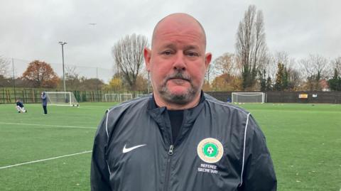 The head and shoulders of Stuart Nixon looking at the camera, with a 4G all-weather green football pitch behind him. He has a beard. he is wearing a black raincoat which is branded with a crest. It is a cloudy day.