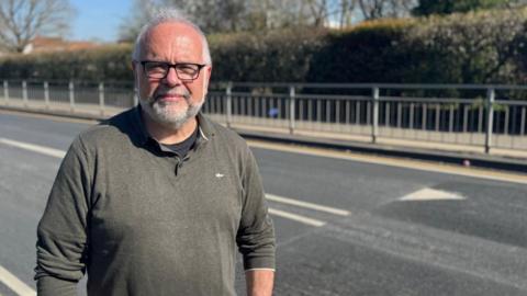 A man wearing a khaki jumper over a black T-shirt looking straight at the camera with a neutral expression. He is standing on a pavement in front of a road with newly installed speed bumps.
