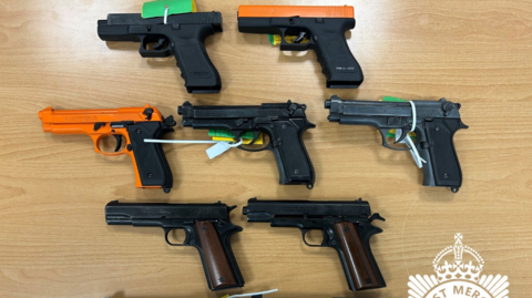 A number of blank firing guns on a wooden surface. Two are at the top, three are in the middle and two are at the bottom near part of the West Mercia Police logo.