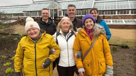 This image shows six people, standing in two lines of three, looking straight at the camera and standing in front of a large greenhouse. There is a tree to their left and outdoor furniture, made of wood, in the background.