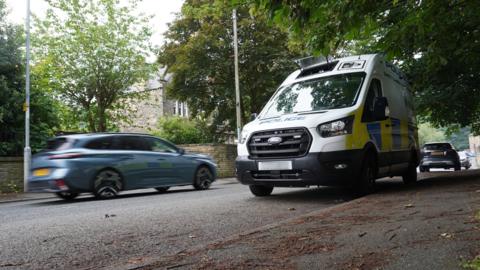 A blurred blue car speeds past a police speed camera van on a residential street.
