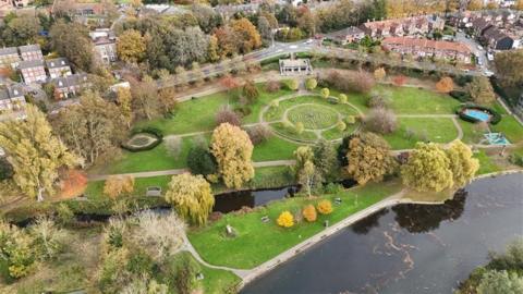 An aerial image of landscaped gardens, mostly laid to lawn with mature trees and pathways. Centre-right is a pathed, circular labyrinth/turf maze in the grass. To the bottom right of the image is the dark water of the River Wensum. To the top of the image, the park is bordered by a road running left to right, with housing and trees beyond.