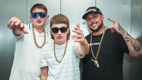 Sam Robinson (Clive), Gareth Kelly and Kane Welsh from Bad Boy Chiller Crew pose, with their fingers doing salute signs. The trio are wearing gold chains around their necks, with Clive and Kane also wearing sunglasses and pale tshirts. Gareth is wearing a black tshirt and cap.