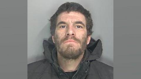 Jason White in a police mug shot. He has curling shortish dark brown hair and a beard. He is wearing a black jacket with its hood pushed down around his neck.