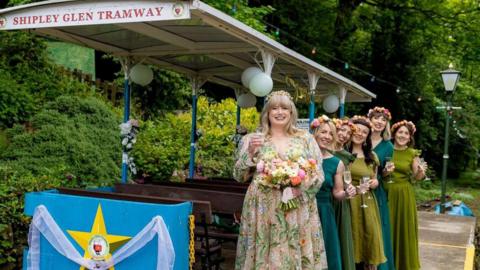 A bride in a floral white dress stands next to a tram with a line of bridesmaids in green dresses behind her
