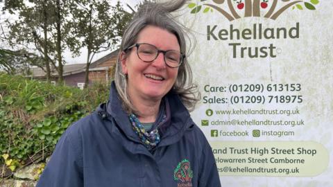 Sally Pyner wearing a blue jacket and stood by a sign for the charity. She has long dark grey hair and glasses. 