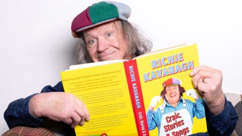 A man with long grey hair holding a yellow and red book, he is smiling at the camera.