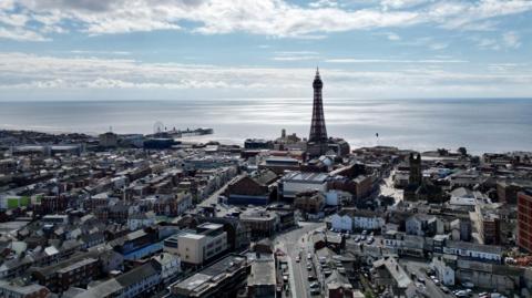 An aerial shot of the town of Blackpool, with the sea in the distance, Blackpool Tower to the left, and streets of houses and shops in the foreground
