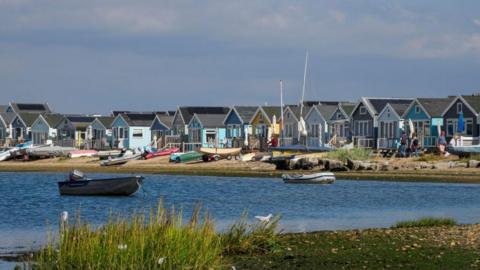 A row of brightly coloured beach huts stand on a sandy beach next to some small dunes. There are picnic benches outside the huts. The sky is blue and it is a sunny day. There is a body of water in front of the beach with some plant life on the near shore. There are several small boats in front of the huts and two in the water.