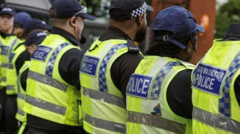 Five police officers stand with their backs to the camera, wearing hi-vis jackets with the blue badges on the back that reads Greater Manchester Police or just the word, Police. 