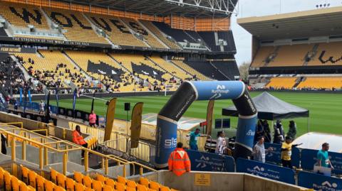 Runners attend the 10k event at Wolverhampton Wanderers Molineux stadium, finish line with RunThrough logo is visible in blue. Security guards wearing neon orange hi-vis are present. Spectators are seen in the distance sitting in the stands