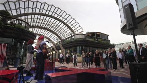 Musicians perform on an outdoor stage in a modern courtyard while a small audience stands watching beneath a large arched glass-and-metal canopy.
