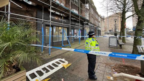 A police officer stands on a street. She is wearing a high-vis yellow jacket, a black hat and black trousers. She is standing on a section of the street that has been cordoned off with blue and white police tape. There is a four-storey building with shops on the ground floor to her left.