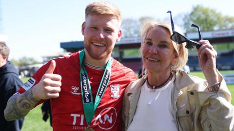 Josh Stones, wearing York City's red shirt with a National League title-winners medal around his neck, smiles and puts his thumbs up while holding his arm around York City owner Julie-Anne Uggla, who is smiling while wearing a white shirt and beige jacket.