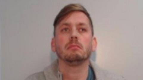 A mugshot of Gregory Bell, who has brown hair swept back to one side and is wearing a grey hoodie. 