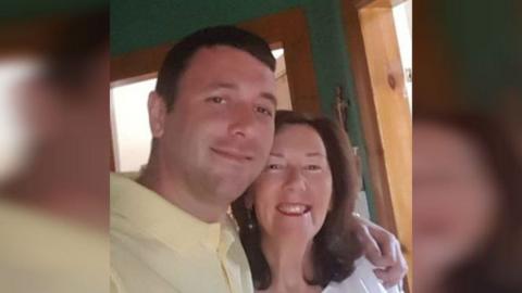 Tony Ferns with his arm around his mum Phyllis Ferns. Tony has short brown hair and wears a yellow polo shirt and smiles. His mum is smaller in height and has shoulder length brown hair and also smiles. They appear to be in a hallway which is painted green.
