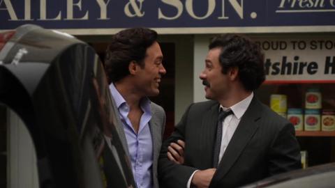 A candid shot of Alex Hassell and Aiden Turner talking to each other on set before filming. Both are dressed smartly in suit jackets, with a car boot up in front of them and a shop behind them