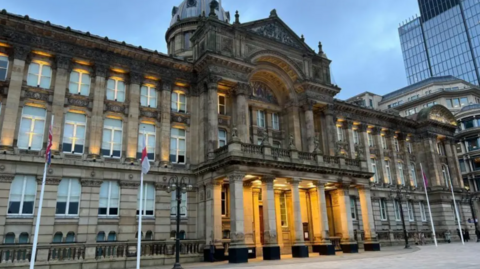 The exterior of the Council House, headquarters of Birmingham City Council. The building has four flagpoles outside it, and comprises of a series of columns and a large archway above the entrance which is lit up by yellow spotlights.
