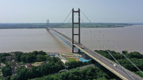 The Humber Bridge stretching across the Humber. The bridge has tall towers and cables supporting it, with vehicles traveling in both directions. Green countryside and scattered houses can be seen on either side of the Humber. The sky is clear and blue.