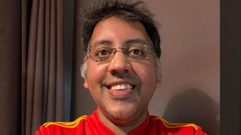 A man with glasses is smiling while taking a selfie image and wearing a red shirt
