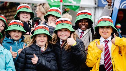 Picture of school children wearing Urdd branded hats and smiling at the camera with their thumbs up. 