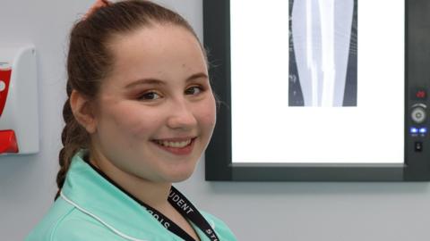 Charley smiles at the camera and is wearing a student lanyard and a paramedic uniform. There is what looks like an X-ray picture of bones behind her.