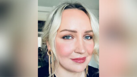 A woman is sitting in a living room. She's smiling into he camera. She has blond hair and is wearing gold earrings.