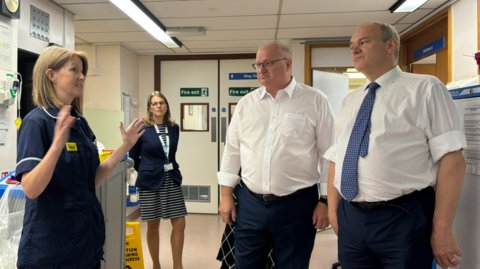 MPs Sir Ed Davey and Ian Roome talk with staff in the Intensive Care Unit at North Devon District Hospital. Both men are wearing navy trousers and white shirts, Sir Ed also wears a blue tie. A female member of hospital staff is talking to them, with her hands gesticulating.