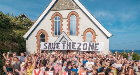 Hundreds of people waving their arms in the air in front of the Methodist chapel with a banner draped across the front of it saying "Save the Zone" 