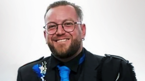 Man in round glasses wearing blue tie and black formal wedding jacket looks at camera.