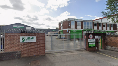 The outside of a recycling centre which is a brown brick building with a green shutter door and white windows, with a large yard. It sits behind a brown brick wall with a silver metal fence. A white sign with green writing reads "LiBatt recycling limited"