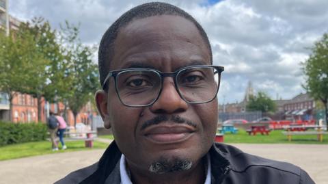 Israel Eguaogie, wearing black glasses and a black jacket, sits in a public square in Belfast