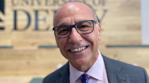 Theo Paphitis, smiling at the camera wearing glasses with a lilac shirt and dark grey blazer, sitting in a University of Derby building