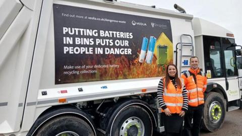 Two staff members in orange hi-vis jackets stand next to a bin lorry showing a sign on the side which reads "putting batteries in bins puts our people in danger".