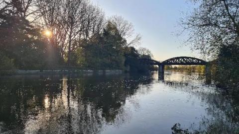 The sun glinting through some tall tress to the left and glistening on a dark river which is in the foreground and snakes off into the distance under a bridge. It is early morning so still a little dark but the sky above is blue.