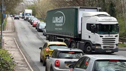 Photo of the A4 Keynsham Bypass congestion. Image shows a lorry in front of a long line of cars. On the other side of the road there is a queue of cars driving by the lorry. The road is surrounded by hedges and trees. 