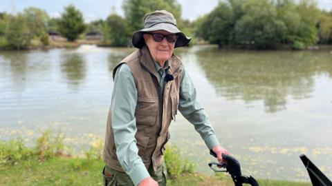 Peter Byers is stood with his rollator walking aid in front of a lake. He is wearing a green fishing hat, sunglasses, a khaki fishing gilet and a grey-green shirt.