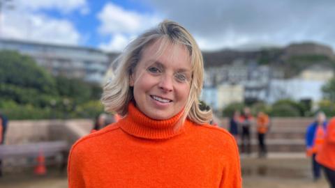 A picture of a woman with blonde hair. She is wearing an orange roll-neck jumper and she is smiling directly at the camera. In the background - out of focus - there are hills with houses on them. There are also a few people in the background.