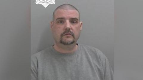Chris Hemming pictured in a police mugshot. He has a blank expression and wears a grey jumper. He has a shaved head, dark stubbly hair and a dark beard.
