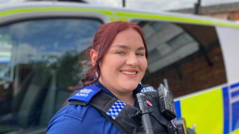 A woman with dark hair smiles toward the camera, they are wearing a blue shirt and a black police stab vest which has a torch and radio attached.
