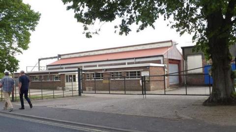 A large factory building can be seen with two men walking past towards the left along the pavement next to the building, which is located behind fencing and a gate. 