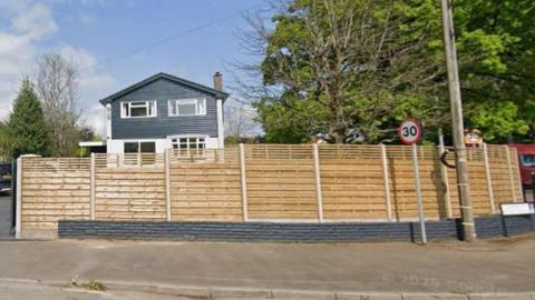 A house on St Lawrence Road, Chepstow. It has a tall timber fence around the edge and a grey slate-style driveway with a dark-coloured SUV parked on it. The house is two storeys and is white on the bottom half with a dark, panel style for the top half. Large trees stand to the right of the house