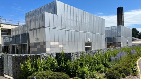 A large grey building, the new critical care unit at the Princess Elizabeth Hospital, with a big black chimney rising over it. 