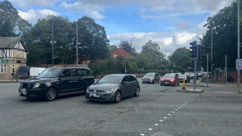 Two lanes of traffic drive along on a busy road at a junction with another road. 