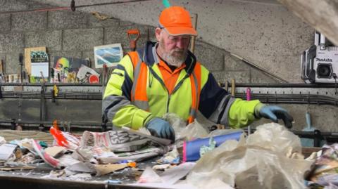A worker at Mayside Recycling sorts through paper waste. He has a beard, is wearing high-vis and a bright orange hat.