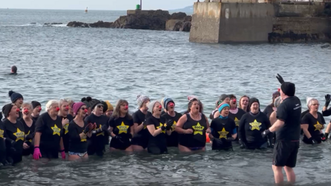 Close up of the choir group in the water. The group are wearing black Rock Choir branded t-shirts. Some members of the choir are wearing woolly hats and plastic red noses. 